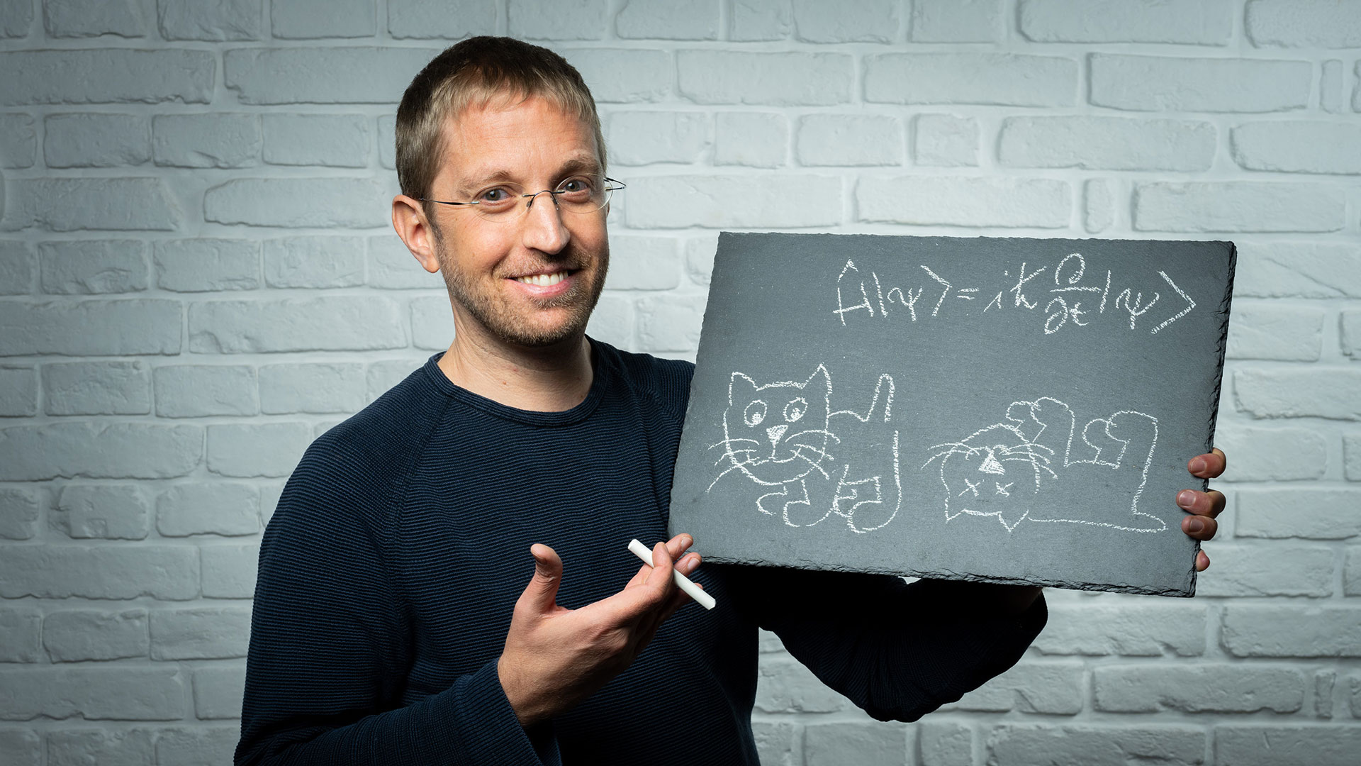 Ein lächelnder Mann mit Brille hält eine Schiefertafel mit Kreidezeichnung. Auf der Tafel ist eine Gleichung aus der Quantenphysik (Wellenfunktion) und zwei Katzen gezeichnet – eine lebendig, eine tot – als Anspielung auf Schrödingers Katze. Der Hintergrund besteht aus einer weißen Backsteinwand.