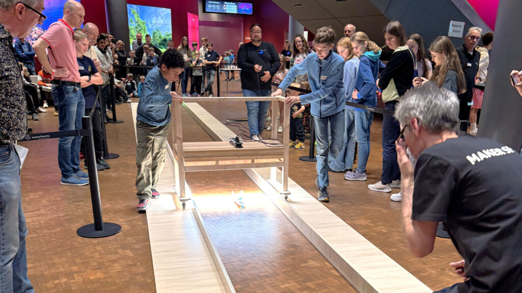 Jugendliche stehen an einer Holzrampe im Innenbereich der experimenta Heilbronn und lassen ihre selbstgebauten Solarmobile auf einer beleuchteten Rennstrecke fahren. Zahlreiche Zuschauer verfolgen gespannt das Rennen beim Solarmobilbauwettbewerb.