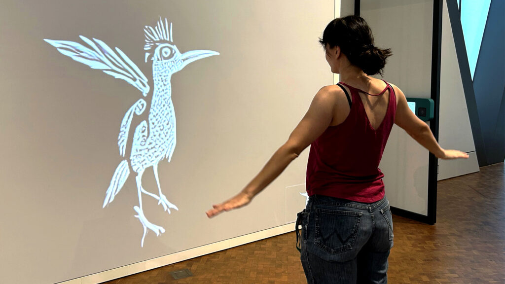 Frau interagiert in der experimenta mit einer KI-Station, die ihre Bewegungen in einen animierten Vogel auf einer Leinwand verwandelt.