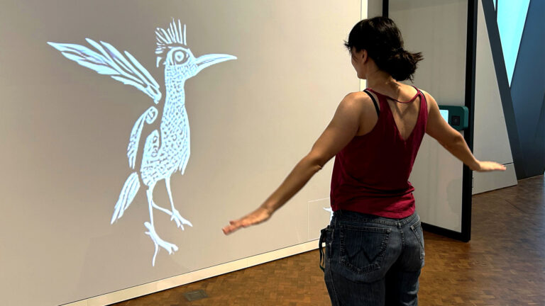 Frau interagiert in der experimenta mit einer KI-Station, die ihre Bewegungen in einen animierten Vogel auf einer Leinwand verwandelt.